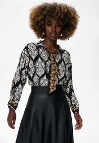 Vrouw met een krullende afro, gekleed in een zwart-wit gestreepte blouse met een contrasterende stropdas en een hoog op de taille zittende zwarte leren rok.