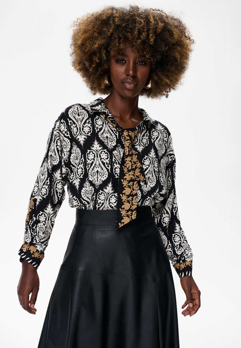 Vrouw met een krullende afro, gekleed in een zwart-wit gestreepte blouse met een contrasterende stropdas en een hoog op de taille zittende zwarte leren rok.