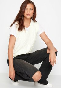 Hvid strikket vest med ribbede kanter, kabelstrik mønster, parret med sorte slidte jeans med knæslidser og hvide flade sko.