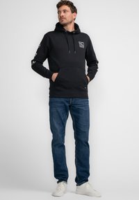 Felpa nera con cappuccio e lacci, tasca frontale, logo sul petto e sulla manica, abbinata a jeans blu e sneaker bianche. Design semplice, in misto cotone.