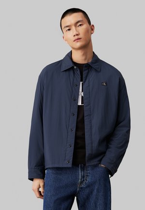 Veste légère bleu marine avec une fermeture à boutons sur le devant, un col pointu et un patch logoté sur la poitrine, associée à un jean en denim foncé.