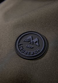 Sort gummipatch med prægede bogstaver "LINDBERGH 1927" og to flag på en olivengrad stofbaggrund med tekstureret finish.