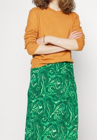 Pomarańczowy sweter z długim rękawem i prążkowanymi mankietami, zestawiony z zieloną spódnicą z wirującym marmurkowym wzorem, tworzącą warstwowy wygląd.