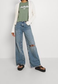 Avskuren grön t-shirt med vit text, vit ribbad cardigan, vida slitna blå jeans och svarta plattform flip-flops.