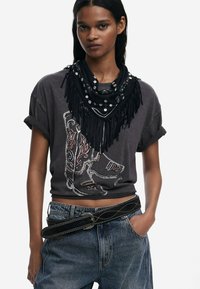 T-shirt graphique noir avec un design de botte de cowboy en blanc et rouge. Porté avec une écharpe noire à franges ornée de clous et une ceinture en cuir.