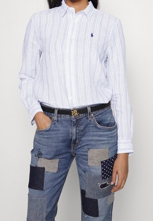 Nainen pukeutunut valkoiseen ja siniseen pystysuoraan raidalliseen napilliseen paitaan ja tilkkutäkkifarkkuihin, käsi taskussa yksiväristä taustaa vasten.