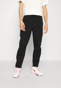 Pantalones jogger negros de corte relajado con cintura elástica y cordón, fabricados con material ligero. Modelo con camisa blanca y zapatillas.