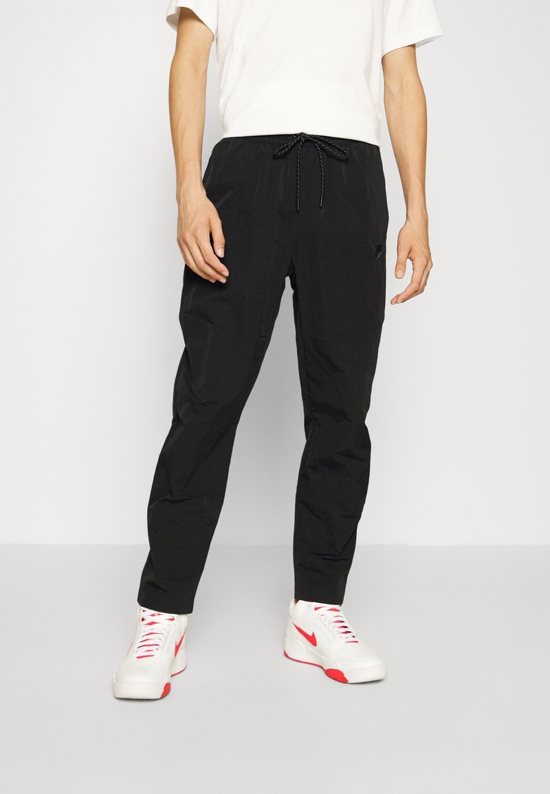Pantalones jogger negros de corte relajado con cintura elástica y cordón, fabricados con material ligero. Modelo con camisa blanca y zapatillas.