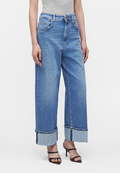 Personne portant un jean bleu clair à jambes larges avec de grands revers et des sandales ouvertes à talons noires, debout devant un fond uni.