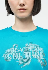 Νεαρός/νεαρή που φοράει μπλε φούτερ με το κείμενο «VERSACE JEANS COUTURE» και διακοσμητικά ασημένια παγιέτες στο μπροστινό μέρος.