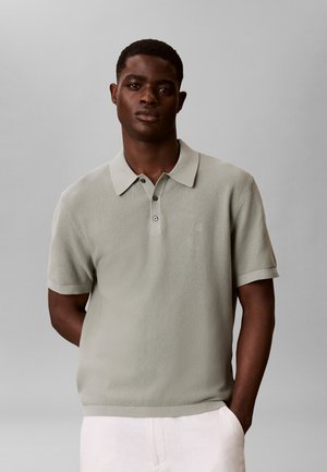 Mand iført en lysegrå tekstureret kortærmet poloshirt med tre knapper, stående med den ene hånd i lommen på hvide bukser mod en ensfarvet baggrund.