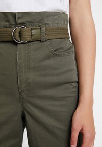 Olivově zelené kalhoty s bočními kapsami, opatřené tkaným opaskem se stříbrnou kovovou sponou. Hladký povrch s viditelnými švy.