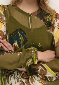 Blusa floreale verde in tessuto trasparente, con colletto abbottonato e maniche lunghe con polsini arricciati che mettono in risalto colori e trame vivaci.