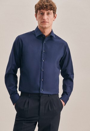 Chemise à manches longues en navy, en tissu lisse, avec un col classique et des boutons contrastants, associée à un pantalon sombre.