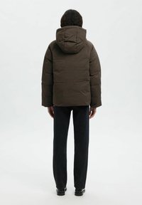 Giacca marrone imbottita con cappuccio, caratterizzata da una forma squadrata, una superficie leggermente lucida e cuciture visibili. Indossata con pantaloni in denim scuri.