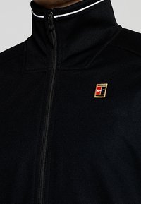 Giacca sportiva nera con zip, colletto alto e un piccolo logo Nike rosso, bianco e oro sul petto.