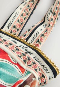 Tracolla di borsa in tessuto con motivi floreali, bordo di perline dorate e testo francese "vacances à la plage" su sfondo crema.