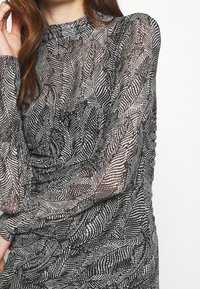 Mustavalkoinen pitkähihainen paita, joka on valmistettu läpikuultavasta kankaasta ja siinä on lehtikuvio. Suunnittelussa on korkea kaulus ja rypytettyjä yksityiskohtia.