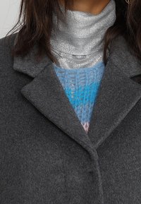 Šedý vlněný kabát s klopovým límcem, vrstvený přes modrý pletený svetr s texturovaným vzorem a pod tím lesklý stříbrný rolák.