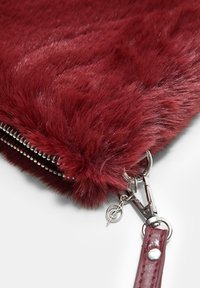 Burgundski faux krzneni clutch s srebrnim zadrge, kovinskimi elementi za ključe in trak. Mehka tekstura z gladkim zaključkom in subtilnim sijajem.