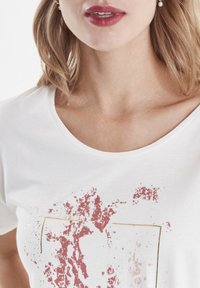 Biała koszulka z okrągłym dekoltem i nadrukiem na przodzie z czerwonymi plamami oraz złotą obwódką. Miękka w dotyku, z krótkimi rękawami.