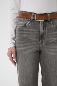 Hallid teksapüksid kõrgema vöökohaga ja sirge lõikega, millel on pruun seemisnahast rihm ja õrnad tuhmumised reitel. Valge pika varrukaga särk paistab.