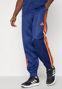 Jūras spēku sporta bikses ar oranžām sānu svītrām, elastīgu jostu un elastīgiem potīšu galiem. Logo atrodas labajā augšējā kājā.