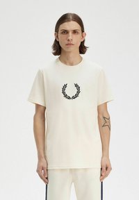 Camiseta corta de manga corta color crema, hecha de algodón, con un bordado de una corona de laurel negra en el pecho y un escote redondo.