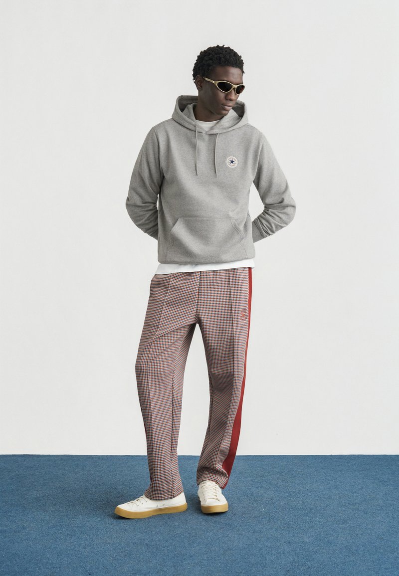 Grå hoodie med kængurulomme og logo, kombineret med ternede bukser med rød sidestribe. Modellen bærer hvide sneakers og solbriller.