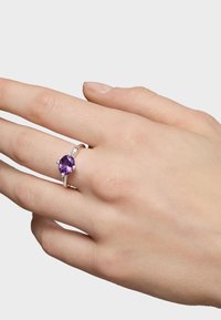 Srebrny pierścionek z okrągłym fioletowym kamieniem i małymi srebrnymi kuleczkami noszony na środkowym palcu prawej ręki na jednolitym tle.