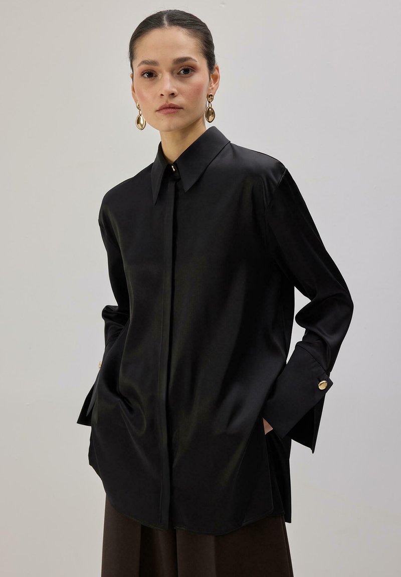 Sötét, hátrafésült hajú nő fekete, túlméretezett szaténingben arany gombos mandzsettákkal és arany csepp alakú fülbevalókkal, kezét a zsebében tartva.