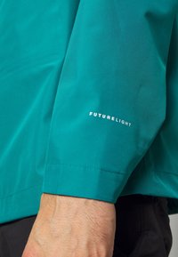 La manche de la veste FUTURELIGHT teal présente une texture lisse et une coupe droite, avec une étiquette sur le poignet affichant le nom de la marque en blanc.
