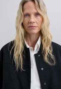 Mujer rubia con ojos azules, vistiendo una camisa blanca abotonada y una chaqueta negra de rayas, mirando hacia adelante frente a un fondo liso.