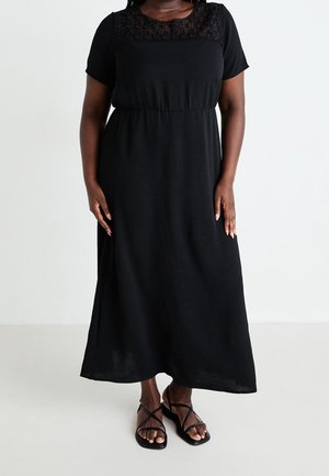 Nainen pukeutunut mustaan lyhythihaiseen maksimekkoon, jossa on pitsiyksityiskohtia rinnan yläosassa, ja mustiin remmillä varustettuihin litteisiin sandaaleihin.