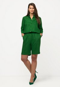 Camicia verde con bottoni, dotata di tasca e maniche lunghe, abbinata a pantaloni corti verdi coordinati. Tessuto liscio, vestibilità rilassata e design elegante.
