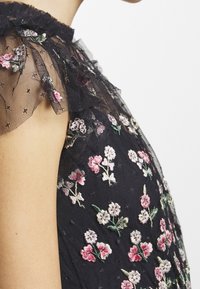 Černé šaty bez rukávů s průhledným volánkovým výstřihem a vyšívanými květinovými vzory v růžové, bílé a zelené barvě na látce.