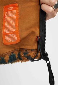 Bruine kledingstuk met een textuurstof en een harige blauwe en bruine zoom. Inclusief een oranje label en een zwarte rits met een koord.