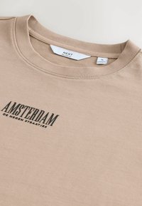 Bežs kokvilnas T-kreklis ar brīvu piegriezumu, uz kura ir melns drukāts attēls "AMSTERDAM de negen straatjes" un apaļa kakla izgriezums.