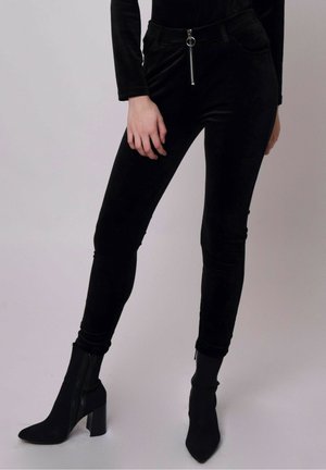 Personne portant un pantalon en velours noir moulant avec fermeture éclair devant et des bottines noires à talons, sur un fond uni.