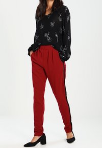 Zwarte blouse met bloemenprint, rode tapered broek met zwarte zijstrepen, gedragen met zwarte flats. Gladde stof, ontspannen pasvorm, lange mouwen.