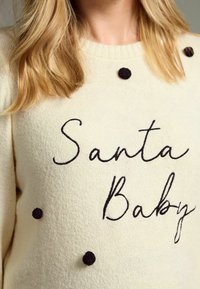 Pull crème avec texte brodé noir "Santa Baby" et trois pompons en feutre noir. Présente un col côtelé et un tissu doux et texturé.