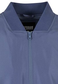 Svijetloplavi bomber jakna s visokim ovratnikom, središnjim patentnim zatvaračem i glatkom teksturom. Sadrži oznaku koja prikazuje "Urban Classics" unutra.