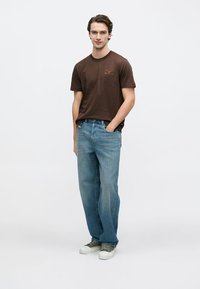 Καφέ κοντομάνικο μπλουζάκι με τύπωμα μπροστά, συνδυασμένο με ανοιχτό μπλε τζιν παντελόνι ίσιας γραμμής και γκρι/άσπρα αθλητικά παπούτσια σε απλό φόντο.