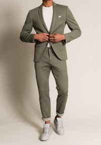 Blazer sartorial verde oliva abbinato a jogger e sneakers bianche. I pantaloni presentano orli arrotolati e il blazer ha una tasca sul petto con un motivo.