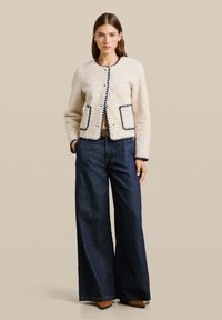 Crème geklede jas met marineblauwe afwerking, voorzien van twee zakken en een ronde halslijn. Gepaard met een wijde donkerdenim jeans en bruine schoenen.