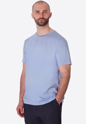 Uomo con barba corta che indossa una camicia azzurro chiaro a maniche corte e pantaloni scuri con una mano in tasca su uno sfondo semplice.