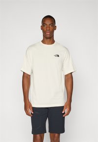 Light beige short-sleeve t-shirt with a round neckline, featuring a small black logo on the left chest and a relaxed fit.