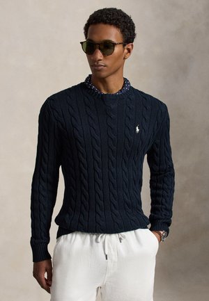 Homme portant un pull torsadé bleu foncé, un pantalon blanc, des lunettes de soleil et une chemise bleue à pois, debout devant un fond neutre.