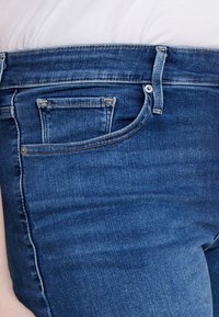 Siniset farkut, joissa on klassinen viiden taskun muotoilu, vetoketjullinen haarakiinnitys ja sävyttynyt ompelu. Materiaali näyttää sileältä ja hieman joustavalta.