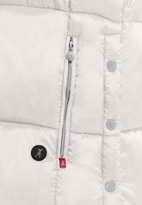 Odcinek jasnobeżowej kurtki puchowej z bocznym zamkiem błyskawicznym, okrągłymi guzikami i małą naszywką z logo psa w czerni i czerwieni. Gładka tekstura.